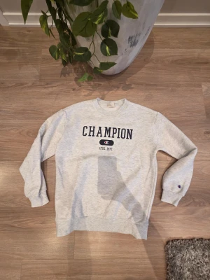 Grå sweatshirt från Champion - Säljer en ljusgrå sweatshirt från Champion med tryckt logga och text på bröstet. Tröjan har rund hals, långa ärmar och ribbade muddar vid ärmslut och nederkant. Perfekt för chill eller sportiga dagar. Klassisk och enkel design med liten Champion-logga på ärmen. OBS! Storleken är XL i barn!✨️