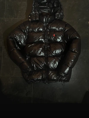Svart pufferjacka från Polo Ralph Lauren - Säljer en svart, glansig pufferjacka från Polo Ralph Lauren med huva och dragkedja framtill. Jackan har en röd Polo-logga på bröstet och är tillverkad i syntetmaterial. Passformen är normal och jackan är perfekt för kalla dagar.