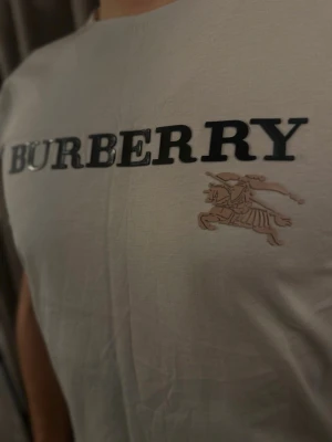 Grey Burberry t-shirt med tryck - Snygg vit t-shirt från Burberry med stort svart logotryck över bröstet och en beige ridande riddare. Klassisk rund hals och korta ärmar. Mjuk bomullskvalitet som känns skön mot huden. Perfekt för dig som gillar stilrena designer och ikoniska märken.