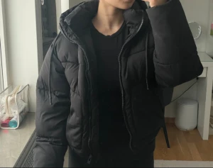 Svart pufferjacka från Pull&Bear - Säljer en svart pufferjacka från Pull&Bear i mycket bra skick. En kort modell som är både varm och stilren, perfekt för kallare dagar. Jackan har en snygg matt dragkedja, luvan är utrustad med justerbara snören för extra komfort och skydd mot kyla. Dessutom finns två praktiska fickor framtill. På insidan kan jackan spännas åt i sidorna för en mer figurnära och anpassad passform.  Jackan är knappt använd. Det finns endast några enstaka lösa trådar, inget som påverkar helheten.