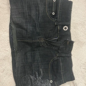 Svart jeanskjol med broderi - Cool svart jeanskjol från Tom Tailor med slitna detaljer, broderi på bakfickan och klassiska jeanssömmar. Kjolen har dragkedja framtill, bälteshällor och rå kant nertill för en edgy look. Perfekt för dig som gillar streetstyle.