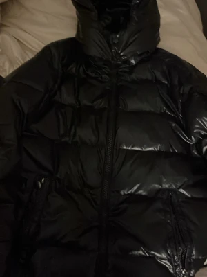 Svart pufferjacka med huva - Säljer en svart pufferjacka med glansig yta och huva. Jackan har dragkedja framtill och två sidofickor med dragkedja. Den är vadderad och har en klassisk pufferlook med breda kanaler. Perfekt för kalla dagar och riktigt snygg stil. Den är från någon hemsida där jag köpte den och är som i nytt sick kan även skicka hur den ser ut på privat 💗
