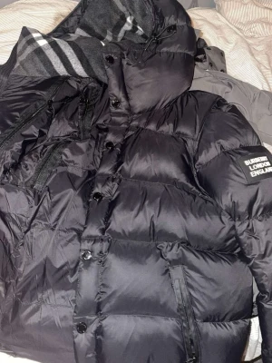 Svart pufferjacka från Burberry - Svart pufferjacka från Burberry med huva, dragkedjor och knappar framtill. Jackan har en patch med Burberry London England på ärmen och flera praktiska fickor med dragkedja. Materialet är glansig polyester och jackan har en quiltad design som ger extra volym. Jackan är nästan helt ny har bara provat den. Den finns i 2 storlekar i S och M.