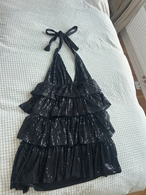Svart glittrig klänning  med knyt - Säljer en svart glittrig klänning med flera lager volanger och knyt i nacken. Toppen är ärmlös och har en djup v-ringning. Perfekt för att sticka ut med paljetter och coolt fall. Passar dig som gillar att synas och vill ha något extra till kvällen.