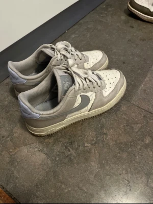 Nike Air Force 1 i grått och vitt - Nike Air Force 1 sneakers i en clean mix av grått och vitt. Klassisk låg modell med snörning, perforerad tå och rund sula. Swoosh-logga i grått på sidan och detaljer i syntetmaterial. 