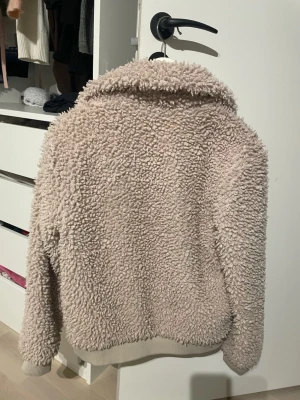 Beige teddyjacka från KappAhl - Supermysig teddyjacka i beige från KappAhl, storlek 170. Jackan har fluffigt material, krage och dragkedja framtill. Perfekt för dig som vill ha en varm och chill look under höst och vinter. Ribbade muddar vid ärmslut och nederkant.
