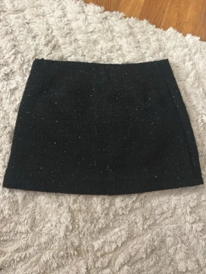 Svart glittrig kortkjol från Faconnable - Säljer en svart glittrig kortkjol från Faconnable i storlek 38. Kjolen har en rak och enkel form med diskret glitter i tyget som ger en snygg touch. Materialet är mjukt och har en fodrad insida för extra komfort. Perfekt att styla med boots eller sneakers.