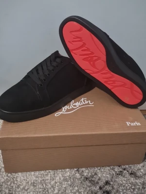 Svarta sneakers från Christian Louboutin - Säljer ett par svarta sneakers från Christian Louboutin med klassisk röd sula och snyggt Louboutin-tryck undertill. Skorna har rund tå, snörning och är tillverkade i mocka med detaljer i syntet. Perfekt för dig som vill ha en lyxig och stilren look.