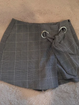 Grå rutiga skorts med knyt - Snygga grå skorts med rutigt mönster och coolt knyt framtill genom två metallringar. Klassisk look med raka ben och diskreta fickor bak. Perfekt för dig som gillar stilrena detaljer och vill ha något som sticker ut lite extra.