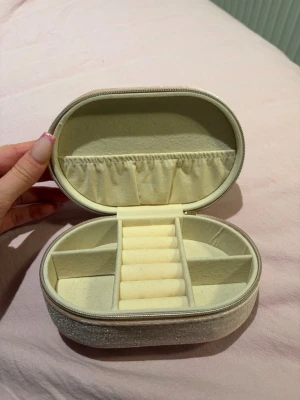 Rosa smyckesväska i sammet - Säljer en oval smyckesväska i rosa sammet med dragkedja runt om. Insidan har flera fack och ringrullar, perfekt för att organisera smycken. Utsidan har en mjuk, glittrig finish som ger en lyxig känsla. Superfin att ha med på resan eller hemma på hyllan.