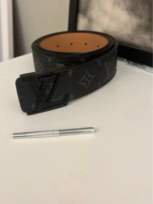 L och v bälte -  skärp med klassiskt monogrammönster i svart och grått. Snygg logga i matt svart metall på spännet. Skärpet är tillverkat i äkta skinn med brunt insida och detaljerad söm. Perfekt accessoar för att lyfta din outfit.