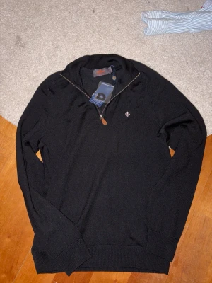 Svart merinoulltröja från Morris - Säljer en svart stickad tröja från Morris i 100% merinoull. Tröjan har en snygg half zip-dragkedja och diskret logga på bröstet. Perfekt för dig som gillar klassisk stil med modern touch. Långärmad och riktigt mjuk. Helt ny med prislappar kvar