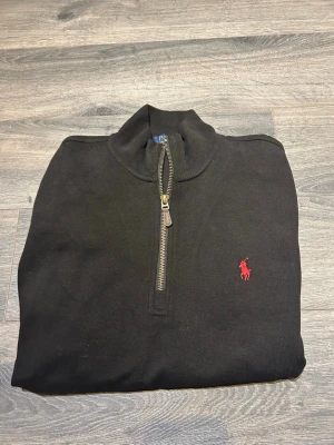 Svart half zip tröja Polo Ralph Lauren - Svart långärmad tröja från Polo Ralph Lauren med klassisk röd logobrodyr på bröstet. Tröjan har en ståkrage och half zip-dragkedja framtill. Materialet är mjuk bomull och passformen är normal. Perfekt för chill dagar och snygg stil.
