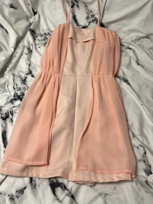 Ljusrosa kort klänning med tunna axelband - Supersöt ljusrosa kort klänning med tunna axelband och lätt draperad framsida. Klänningen har ett flowigt lager i chiffongliknande material och är ärmlös. Perfekt för varma dagar och har en feminin, enkel look. Klänningen har även inbyggda shorts vilket är väldigt praktiskt. 