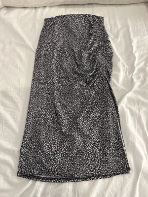 Blommig långkjol från H&M Divided XS - Säljer en svart långkjol med små blommor i rosa och vitt från H&M Divided, storlek XS. Kjolen har en snygg slits på sidan och är gjord i mjuk polyester med lite stretch. Perfekt för dig som gillar blommigt och vill ha en skön kjol med cool detalj.