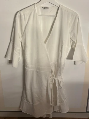 Vit omlottklänning från Bubbleroom - Snygg vit omlottklänning från Bubbleroom i storlek 40. Klänningen har trekvartsärmar med volang, v-ringad hals och knyt i midjan. Materialet känns mjukt och lätt, perfekt för en fräsch look. Enkel och stilren design som är lätt att matcha med accessoarer.