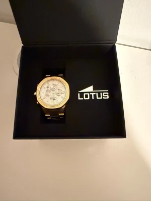 Guldig klocka från Lotus - Guldig klocka från Lotus med vit urtavla. Endast buren en gång, annars i perfekt skick. Klockglasmaterial: Mineralglas, Boett: ädelstpl, Armbandsmaterial: ädelstål, medföljer orginal boxen och en extra del, ifall man vill förstora/förminska