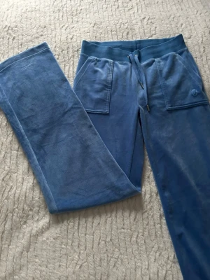 Blå mjukisbyxor med snörning - Säljer ett par blå mjukisbyxor från Juicy Couture med bred resår och snörning i midjan. Byxorna har raka ben, stora fickor fram och en broderad JC-logga på ena fickan. Materialet är mjukt och skönt velour som ger en chill vibe.