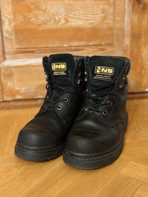 Svarta Northbrook boots strl 39 - Säljer ett par svarta boots från Northbrook i storlek 39. Skorna har robust snörning, rund tå och grov platt sula. Ovandelen är i skinn och resten i syntetmaterial. Guldiga detaljer på etiketten och bak på skaftet. Perfekta för dig som gillar chunky och slitstark stil. Kan behöva nya snören annars är de super fina. 