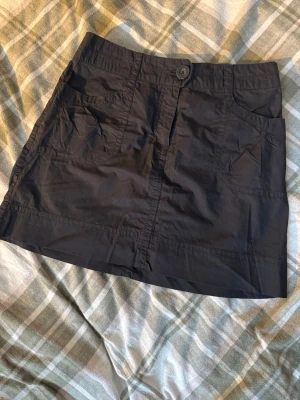 Svart kortkjol från H&M, strl 36 - Svart kortkjol från H&M i storlek 36. Kjolen har praktiska fickor både fram och bak med knappdetaljer och stängs med knapp och dragkedja framtill. Tillverkad i lätt bomullsmaterial som känns skönt mot huden. Perfekt för dig som gillar enkel och stilren design. 36 cm rakt över 