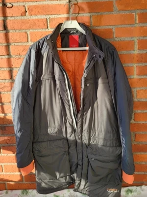 Grå lång parkajacka från J.Lindeberg L - Parkajacka från J.Lindeberg i mörkgrått med orange quiltat foder. Jackan har flera stora fickor, dragkedja och knappar framtill samt hög krage. Materialet är polyester och jackan är perfekt för kyliga dagar. Snygg och funktionell design med coola detaljer.