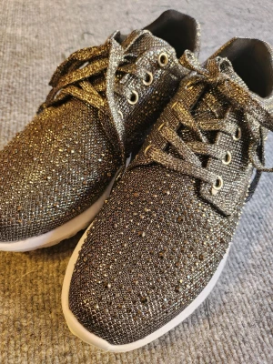 Glittriga sneakers med vita sulor - Säljer ett par snygga sneakers med glittrigt tyg och vita sulor. Skorna har snörning och detaljer i metallic på hälen. Perfekta för dig som vill sticka ut med lite extra bling på fötterna. Skorna har rund tå och är tillverkade i syntetmaterial.