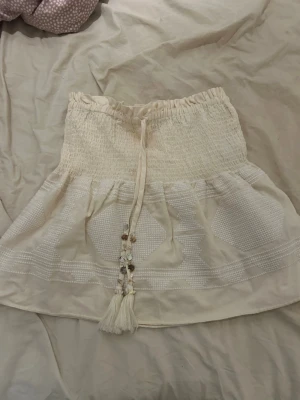   Zara ljus kjol perfekt till sommar – S -  Säljer en kort volangkjol från Zara i storlek S. Färgen är beige/cremevit och ger en ljus, strandig sommarkänsla.  Kjolen har två snören framtill med små dekorativa stenar och tofsar, vilket ger en fin detalj utan att det tar över. Lätt, luftig och bekväm – perfekt till sommar, semester, strand eller festival. Passar bra till linne, topp eller bikinitopp.  🧺 Skick: mycket bra 👗 Storlek: S 🏷️ Märke: Zara 