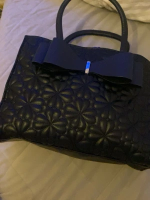 Svart handväska med rosett och blommönster - Säljer en svart handväska med quiltat blommönster och en stor rosett framtill med silverdetalj. Väskan har två handtag och dragkedja upptill. Perfekt storlek för att få plats med allt viktigt och har en riktigt snygg design.