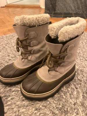 Sorel Caribou vinterboots strl 38 - Säljer ett par Sorel Caribou vinterboots i beige och mörkbrunt med varm ullfoder och snörning. Bootsens ovandel är i skinn och nederdelen i vattentät gummi, perfekt för kalla och blöta dagar. Grov sula för extra grepp och komfort. Bra passform 