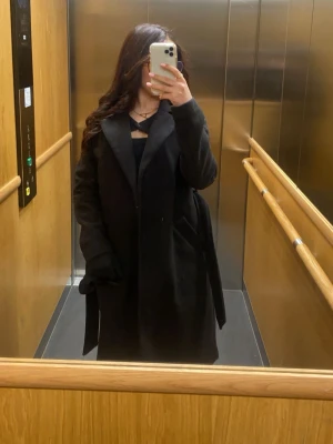 Svart kappa med bälte - Säljer en svart lång kappa med bälte i midjan och bred krage. Jackan har en klassisk och stilren look, perfekt för dig som gillar en enkel men snygg stil. Långa ärmar och djupa fickor på sidorna. Passar till både chill och uppklädda outfits.