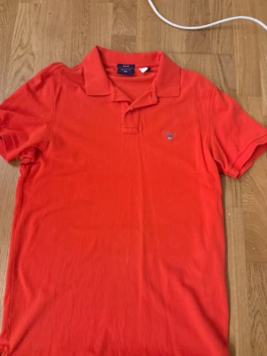 orange pikétröja från GANT, storlek M - Säljer en snygg orange pikétröja från GANT i klassisk modell med krage och korta ärmar. Tröjan har broderad logga på bröstet och är gjord i mjuk bomull som känns skön mot huden. Perfekt för dig som gillar stilrena och sportiga plagg.