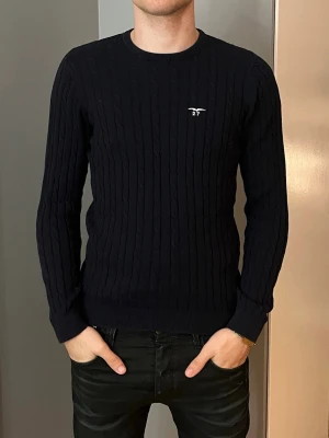 Mörkblå kabelstickad tröja  - Säljer en mörkblå kabelstickad tröja med rund halsringning och diskret logga på bröstet. Tröjan har en klassisk passform och långa ärmar, perfekt för lager-på-lager under kyliga dagar.