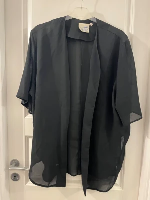 Svart kofta från Airetex Classic - Säljer en svart, tunn och luftig kofta från Airetex Classic. Koftan har öppen front, korta ärmar och är tillverkad i ett lätt syntetmaterial som ger en skön känsla. Perfekt att slänga över en topp för en chill look.