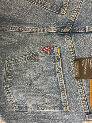 Levi's 501 ljusblå raka jeans - Klassiska Levi's 501 jeans i ljusblå tvätt med raka ben och knappgylf. Jeansen har fem fickor, gul kontrastsöm och den ikoniska röda Levi's-lappen på bakfickan. Tillverkade i slitstarkt bomullsjeansmaterial, perfekta för en avslappnad look.