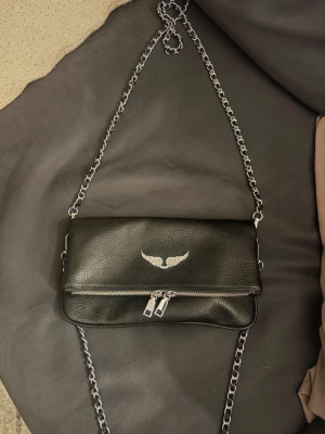 Svart axelväska från Zadig & Voltaire - Säljer en svart axelväska från Zadig & Voltaire i skinn med silverfärgad kedja och detaljer. Väskan har ett snyggt emblem med vingar framtill, dubbla dragkedjor och magnetknappar. Perfekt storlek för det viktigaste och riktigt cool design.