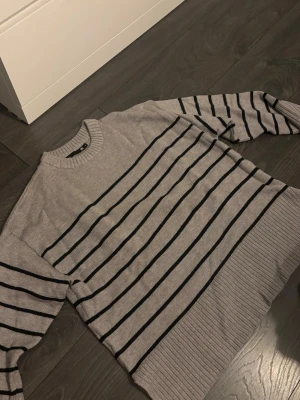 Grå randig stickad tröja 157 - Snygg grå stickad tröja från 157 i storlek S. Tröjan har svarta horisontella ränder och ribbade muddar vid hals, ärmslut och nederkant. Perfekt för dig som gillar enkel men stilren design med lite edge. Använd en gång🌸