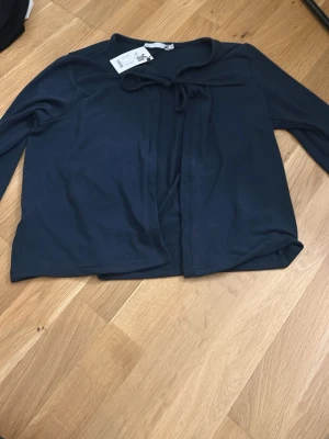 Mörkblå kofta med knyt lager 157 - Mörkblå kofta från lager i mjukt material. Koftan har långa ärmar och en söt knytning framtill vid halsen. Enkel och stilren design som passar perfekt till lager på lager under höst och vår.