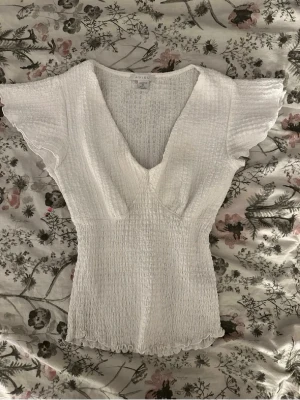 Vit smockad blus från Amisu - Supersöt vit blus  med smockad struktur och v-ringning. Blusen har korta, volangiga ärmar och lätt rynkad kant nedtill. Perfekt till jeans eller kjol för en fräsch look. Materialet känns mjukt och stretchigt.