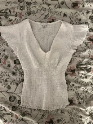 Vit blus - Säljer en vit blus med v-ringad hals och korta volangärmar. Toppen har en smockad, ribbad struktur och är figurnära med lätt rynkad nederkant. .