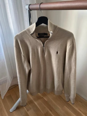 Beige half zip tröja Polo Ralph Lauren - Säljer en beige långärmad tröja från Polo Ralph Lauren i mjuk pima bomull. Tröjan har half zip-dragkedja, ribbade muddar med mörkblå kant och klassisk broderad logga på bröstet. Perfekt för lager på lager under höst och vår.