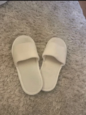 Vita tofflor i mjukt material - Säljer ett par vita tofflor med öppen tå och bred rem över foten. De är gjorda i ett mjukt tygmaterial och har platt sula, perfekta att glida i hemma. Enkla och stilrena för en chill vibe.