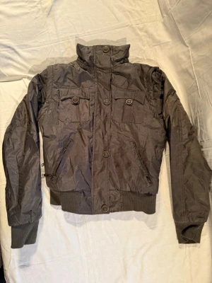 Svart bomberjacka från Lee Cooper - Snygg svart bomberjacka från Lee Cooper med högt krage, ribbade muddar och midja. Jackan har två bröstfickor med knappdetaljer och två sidofickor med dragkedja. Materialet är polyester och insidan har mjuk fuskpäls för extra komfort.