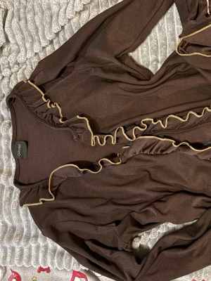 Brun kofta med volang från Catwalk - Säljer en brun kofta från Catwalk med långa ärmar och snygga volangdetaljer längs kanten. Koftan har en guldfärgad kant och knytning framtill. Perfekt att slänga över en topp för en chill och trendig look.