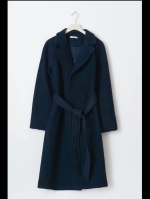 Mörkblå kappa med bälte - Snygg mörkblå kappa från Arket med klassisk krage och knytskärp i midjan. Lång modell med raka ärmar och stilren design. Perfekt för dig som vill ha en tidlös och clean look. Materialet är ullblandning som håller dig varm under kyliga dagar.Säljs inte längre 