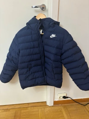 Mörkblå pufferjacka från Nike - Säljer en mörkblå pufferjacka från Nike helt ny, med huva och dragkedja framtill. Jackan har två fickor på sidorna och klassisk Nike-logga på bröstet. Perfekt för kyliga dagar och riktigt skön att bära.