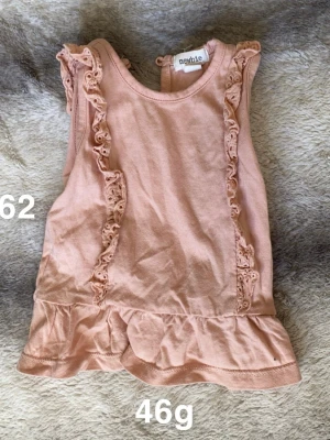 Aprikos ärmlös topp från Newbie - Säljer en söt aprikos ärmlös topp från Newbie med volangdetaljer längs axlar och fram samt en bred volangkant nedtill. Toppen är gjord i mjuk bomull och har en romantisk look som passar perfekt till sommaren.🌸 Fint använt skick. Storlek 62