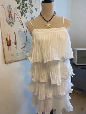 Vit volangklänning student  - Aldrig använd och inga defekter! Köpt vintage för 800kr därav priset! Supersöt vit klänning med tunna axelband och flera lager volanger. Klänningen har ett plisserat tyg som ger en luftig och flowy känsla. Perfekt för sommarens alla tillfällen och ger en riktigt fräsch vibe. 