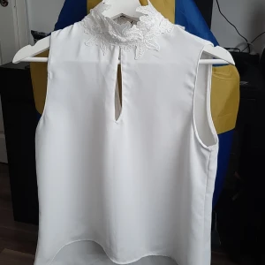 Vit blus med spetskrage och pärlor från Zara  - Supersöt vit blus med hög spetskrage dekorerad med broderade blommor och små pärlor. Ärmlös modell med lätt A-linjeform och snygg öppning framtill under kragen. Perfekt för dig som vill ha en elegant och unik topp med fina detaljer.