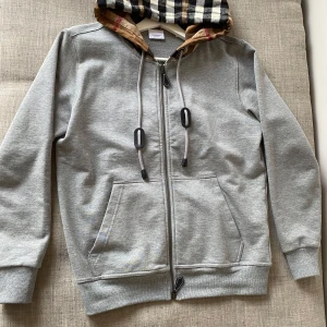 Grå hoodie från Burberry - Tja! Säljer en stilren grå hoodie från Burberry med dragkedja och klassiskt rutigt mönster i huvan. Tröjan har långa ärmar och praktiska fickor framtill. Perfekt för en avslappnad och trendig look.