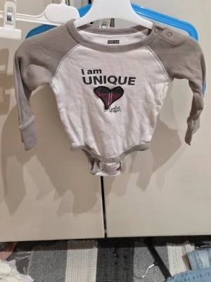 Vit och grå body från Lundmyr - Säljer en söt body från Lundmyr of Sweden i vitt med grå ärmar och kantband. Framsidan har ett tryck med texten 'I am UNIQUE' och ett hjärta i rutig design. Bodyn är tillverkad i mjuk bomull och har praktiska tryckknappar vid axeln och i grenen.